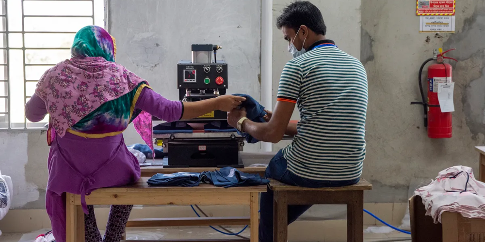 Two garment workers handling denim in a small workshop, representing textile sourcing, labor, and fashion manufacturing