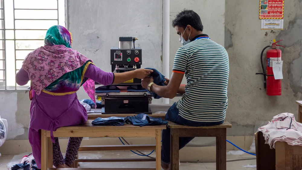 Two garment workers handling denim in a small workshop, representing textile sourcing, labor, and fashion manufacturing