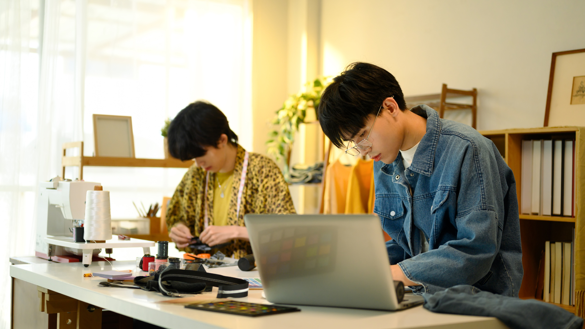 Two fashion designers working in a bright studio. One is focused on his laptop, reviewing the production calendar, while the other works on fabric pieces and sewing in the background.