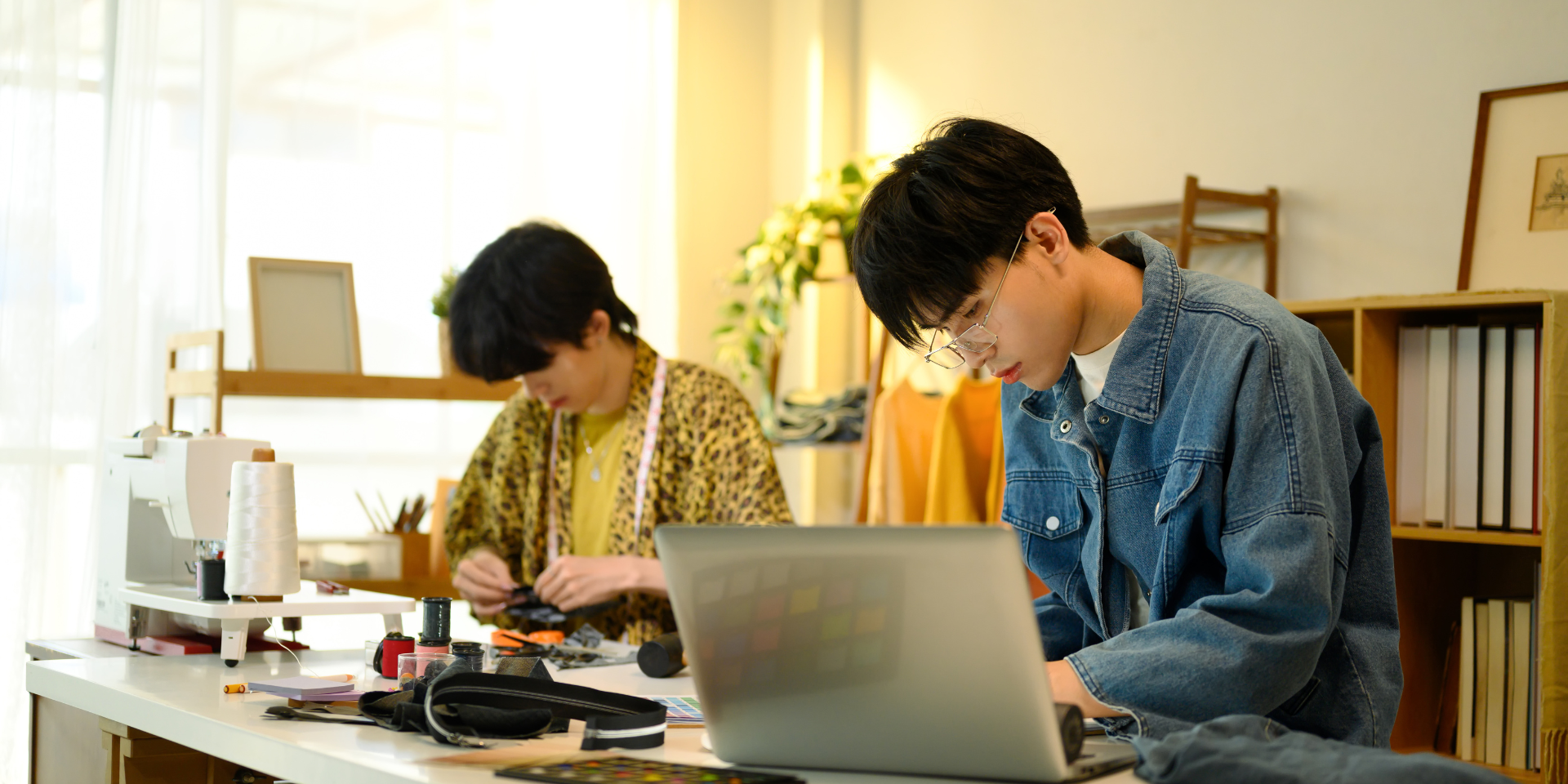 Two fashion designers working in a bright studio. One is focused on his laptop, reviewing the production calendar, while the other works on fabric pieces and sewing in the background.