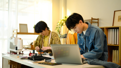 Two fashion designers working in a bright studio. One is focused on his laptop, reviewing the production calendar, while the other works on fabric pieces and sewing in the background.