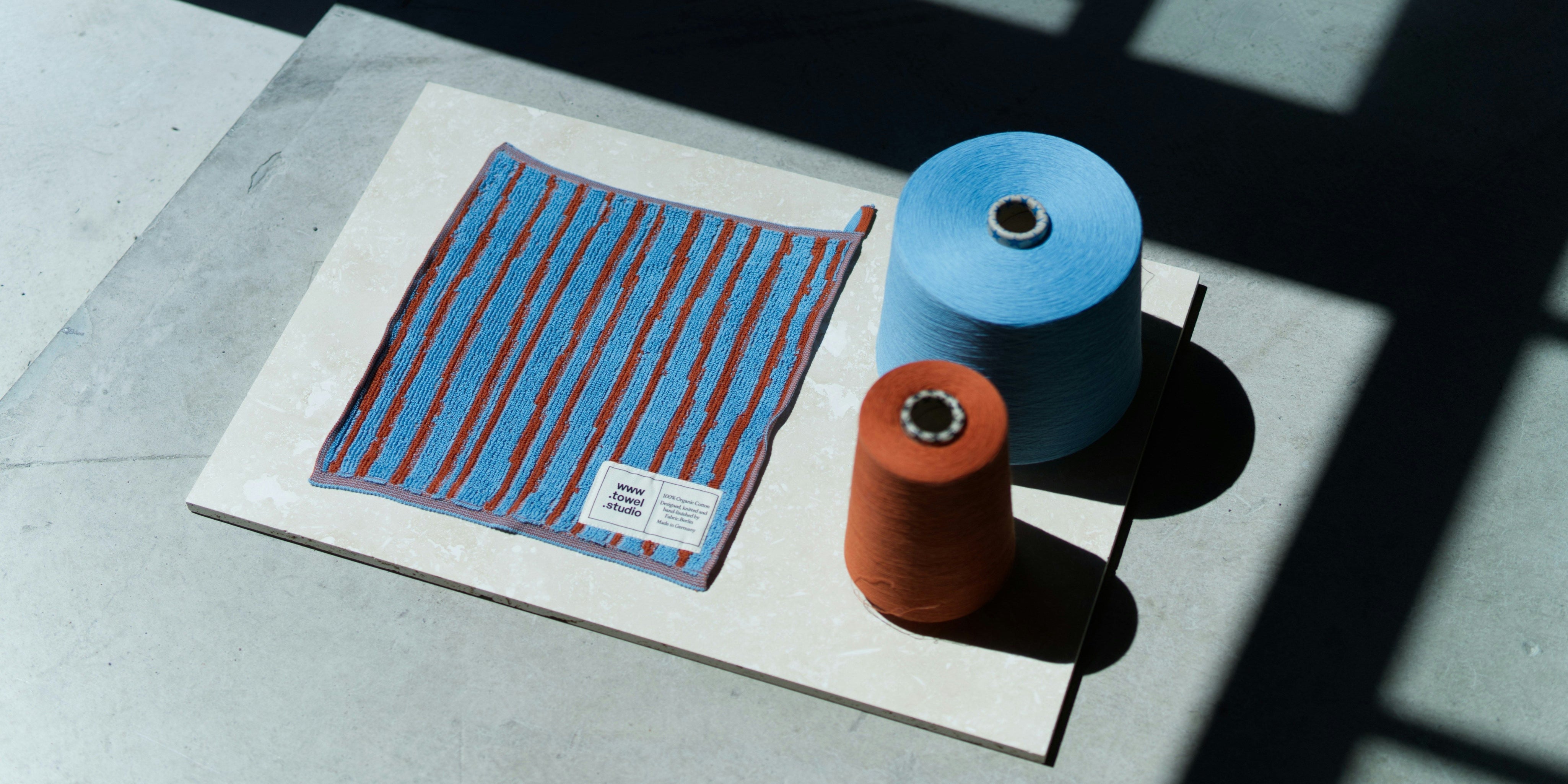 Flat lay of a blue and red striped textile sample displayed alongside two yarn cones, one blue and one rust-orange, on a light stone surface under strong natural light and shadows.