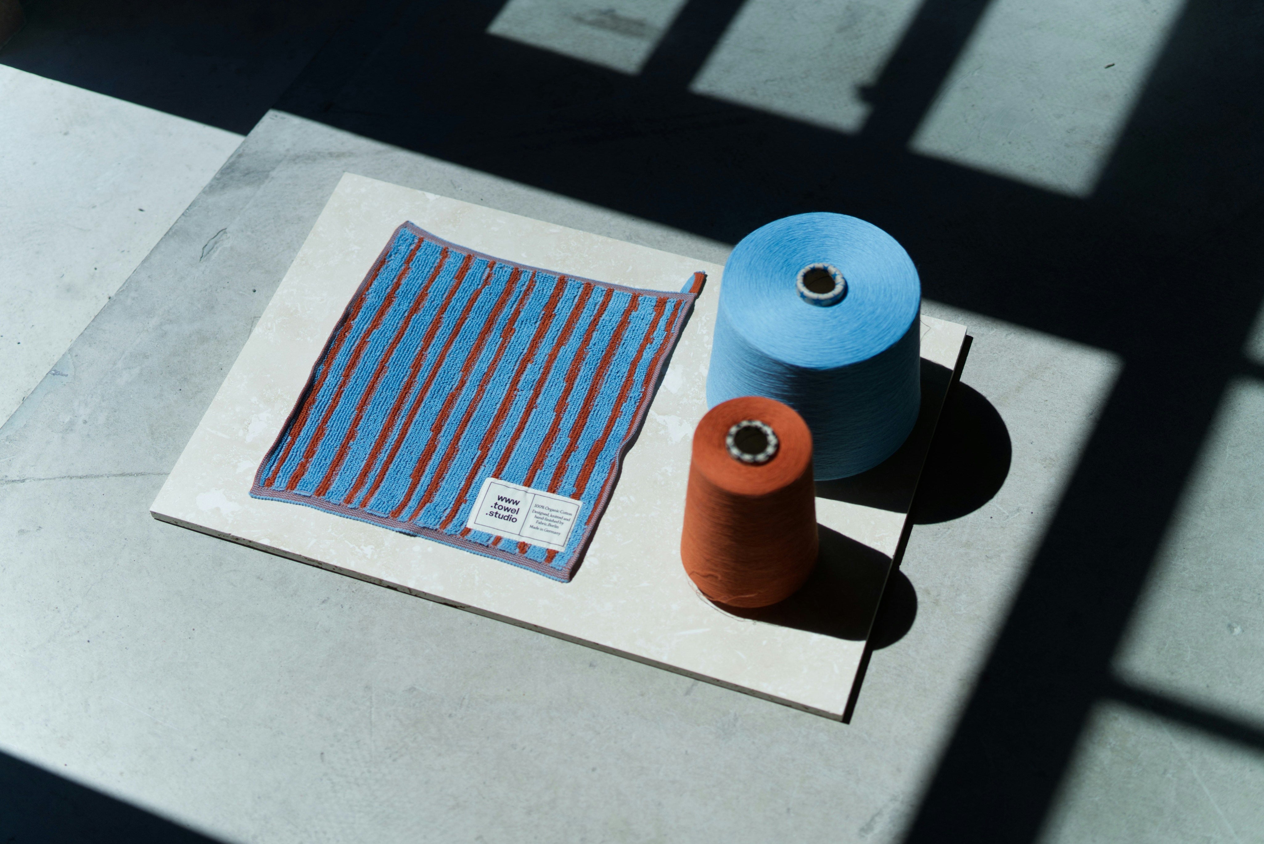 Flat lay of a blue and red striped textile sample displayed alongside two yarn cones, one blue and one rust-orange, on a light stone surface under strong natural light and shadows.