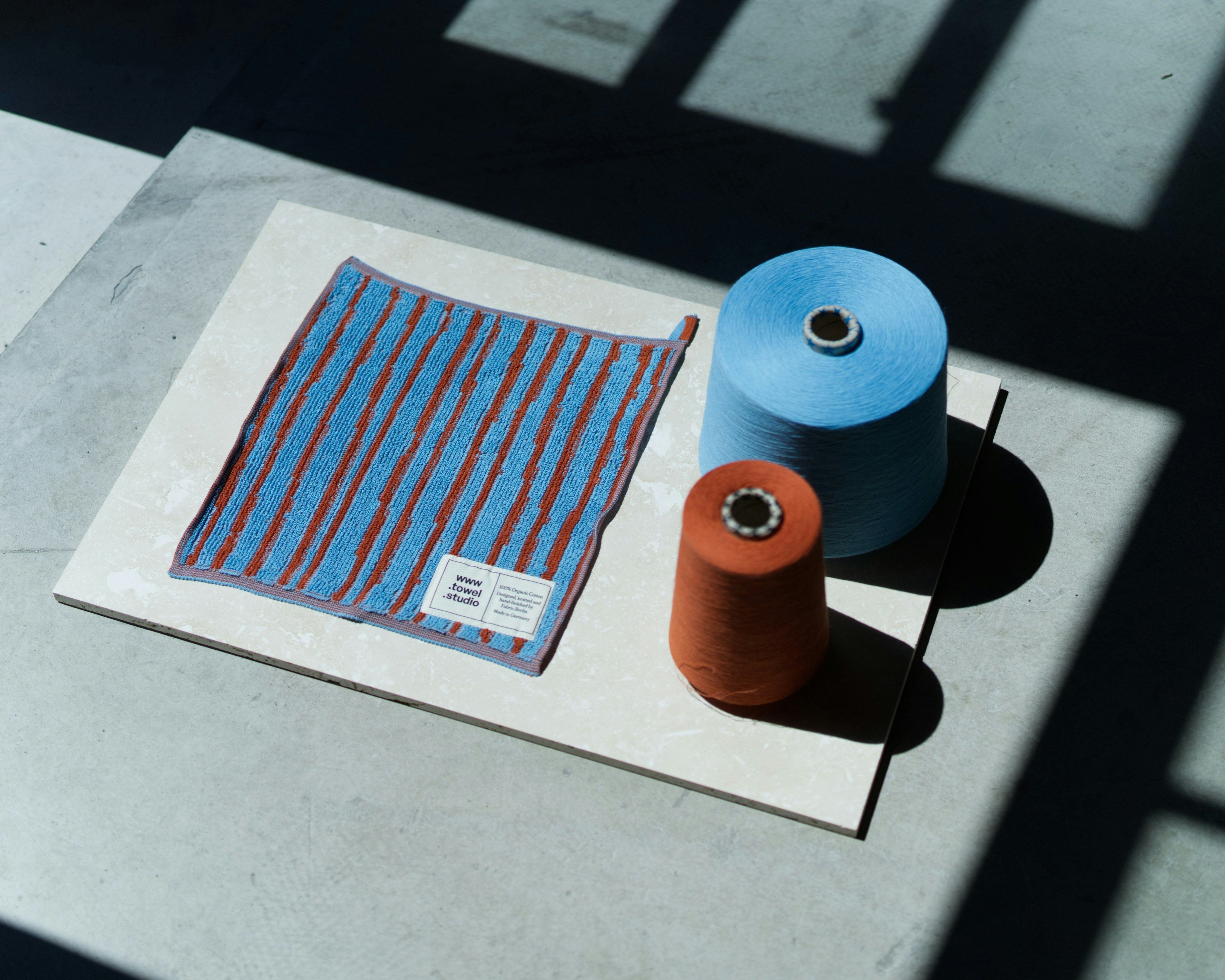 Flat lay of a blue and red striped textile sample displayed alongside two yarn cones, one blue and one rust-orange, on a light stone surface under strong natural light and shadows.